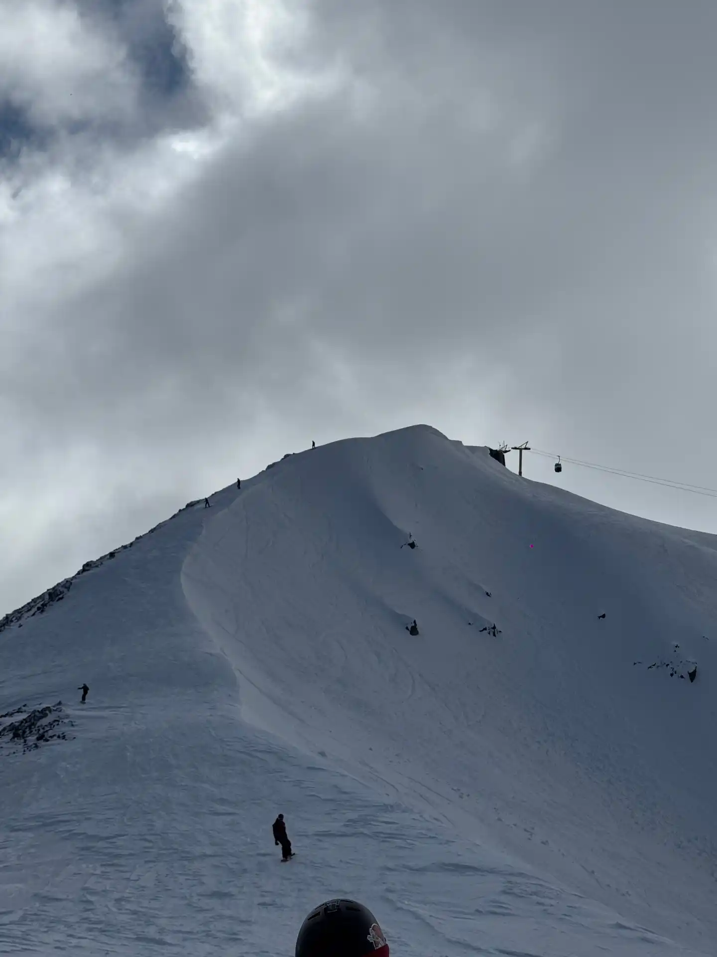 Skiers on a steep snowy bowl at the summit of Cerro Bayo with chairlift and dramatic clouds