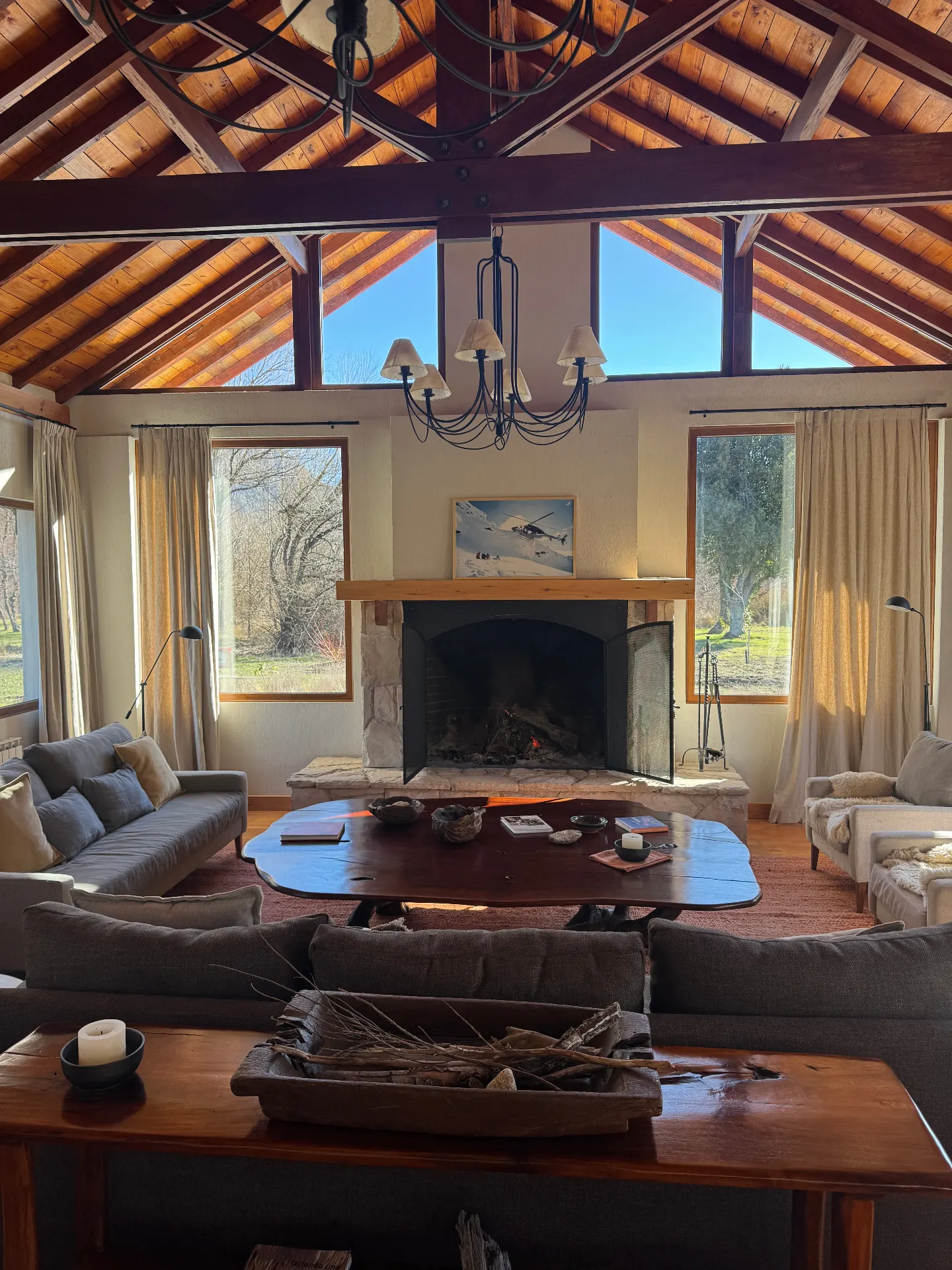 Patagonian ski lodge living room with vaulted wood-beam ceiling, stone fireplace and floor-to-ceiling windows looking onto the valley