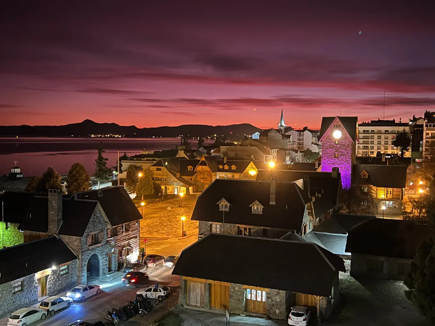 Bariloche at dusk, with dark wooden and stone buildings lit by warm street lights above the lake under an orange and purple sky
