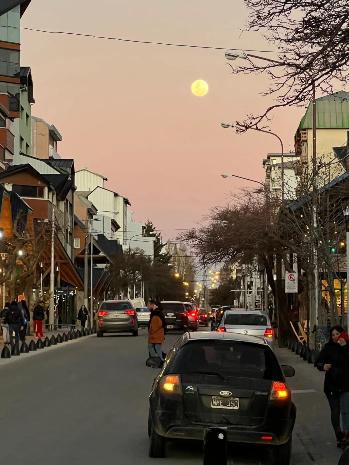 Traffic and pedestrians on a downtown Bariloche street at dusk, with a full moon rising above the shops and apartment blocks