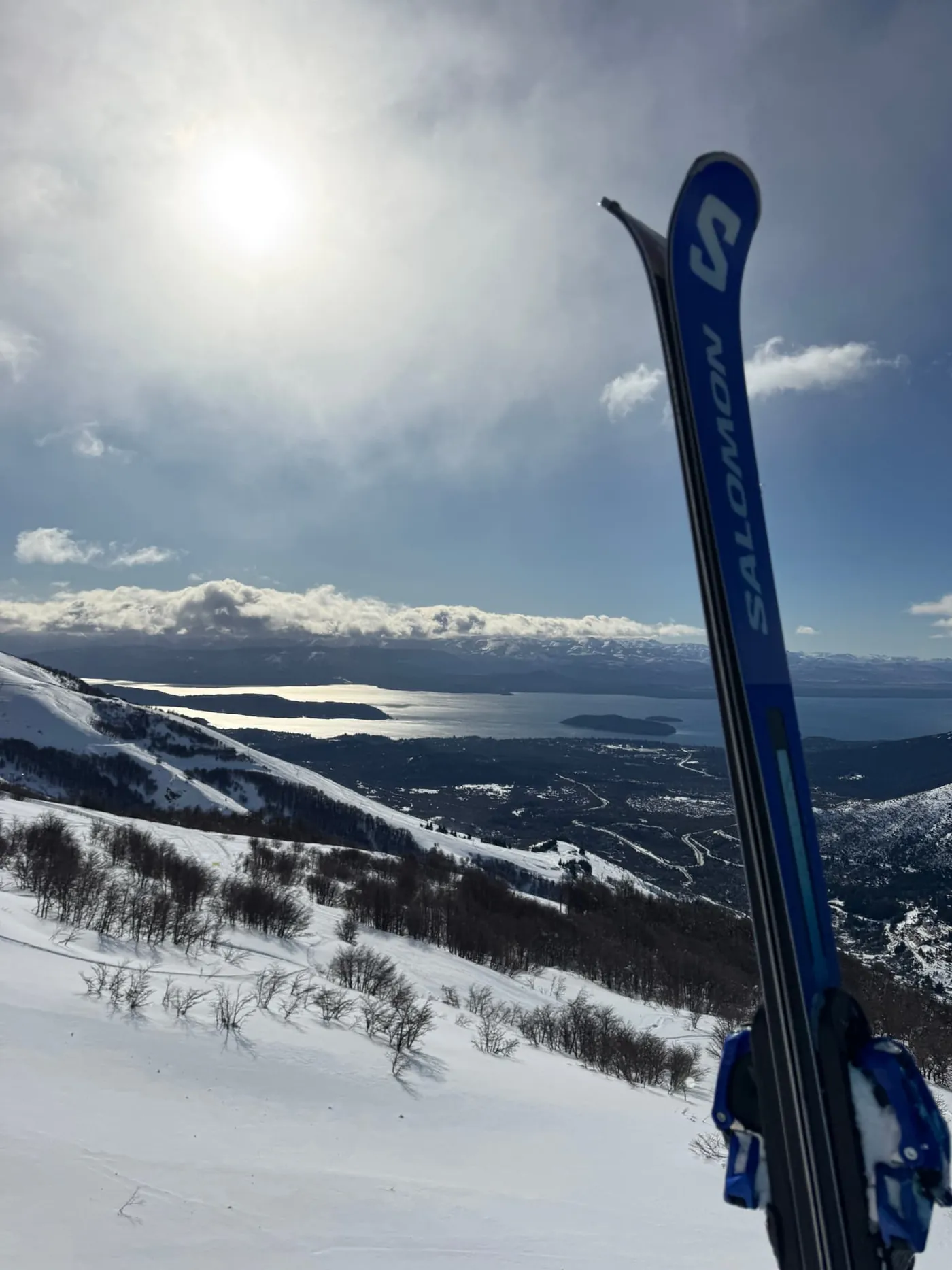 Blue Salomon skis standing upright in the snow at Cerro Catedral, with Lago Nahuel Huapi and the Bariloche valley below