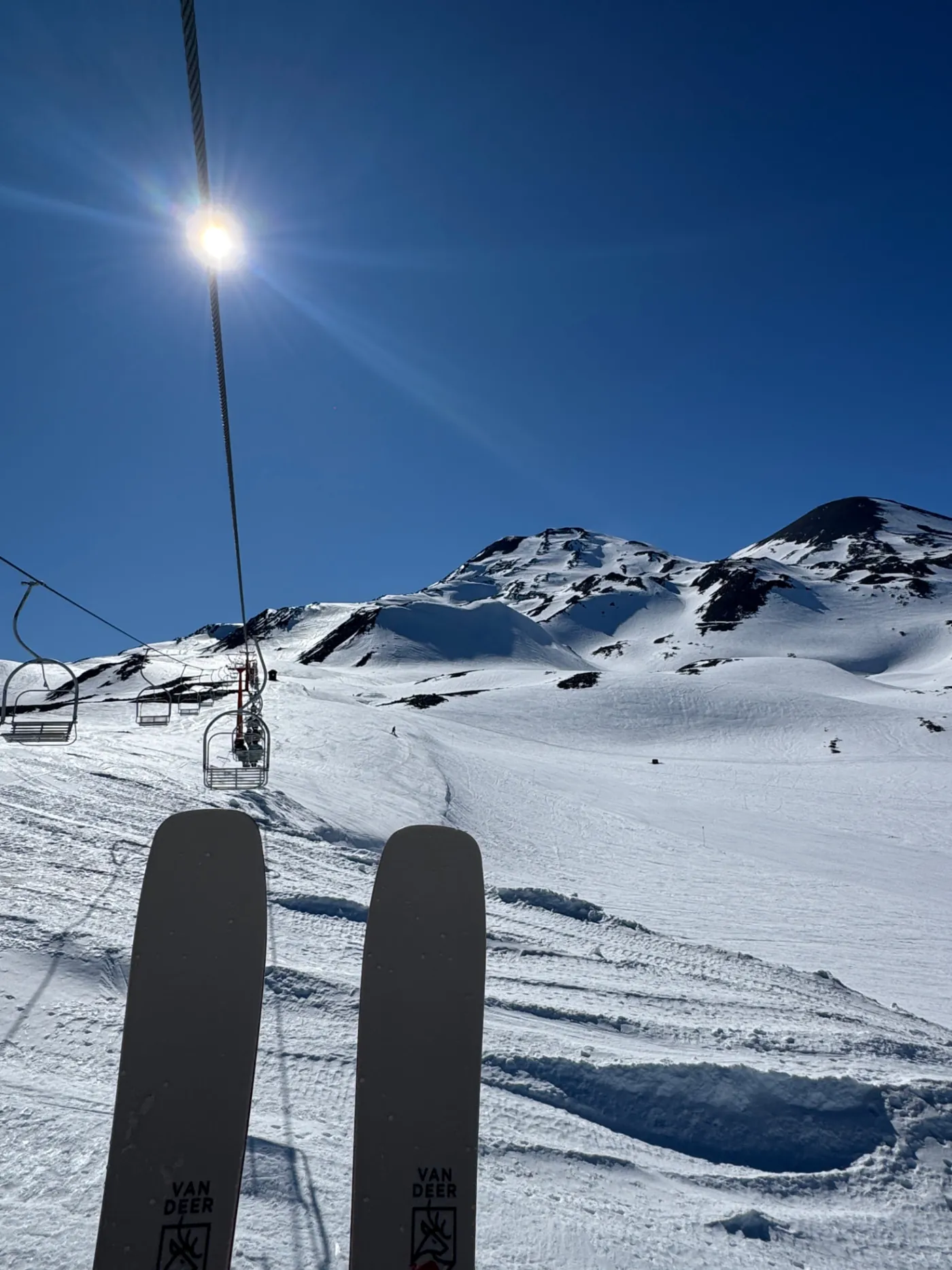 Ski tips seen from a chairlift, pointing up a wide sunlit slope toward a snow covered summit under a deep blue sky
