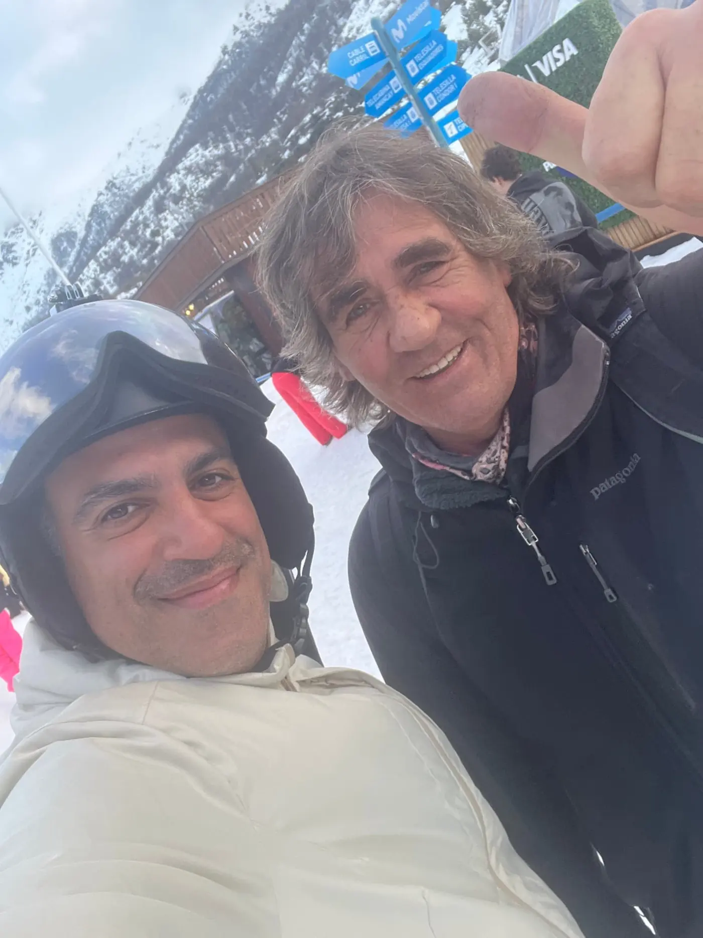 Two men smiling for a selfie in front of a wooden ski resort building and blue trail signs, one wearing a ski helmet and goggles