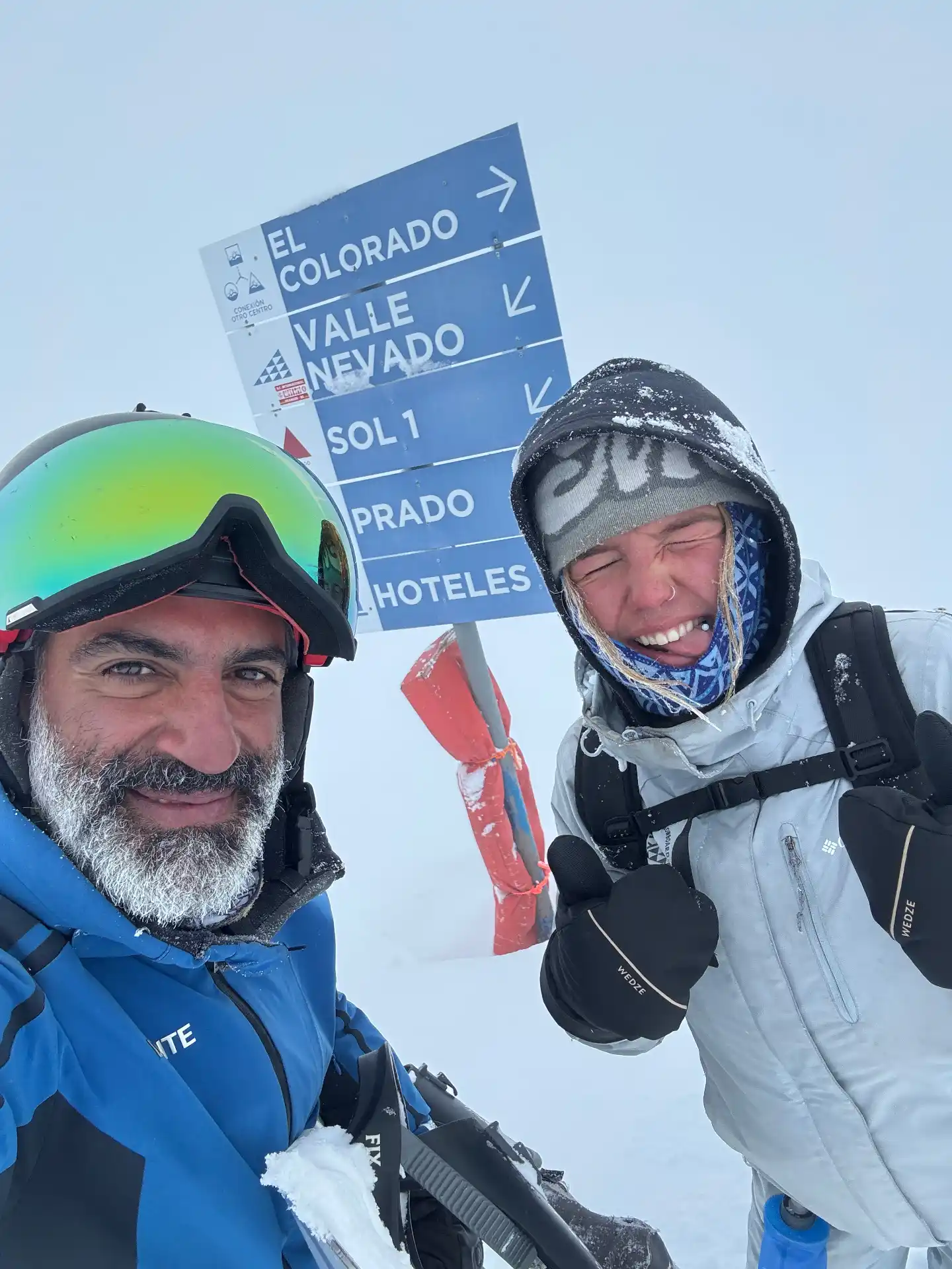Two skiers bundled up in a snowstorm at an El Colorado / Valle Nevado / Sol / Prado trail sign