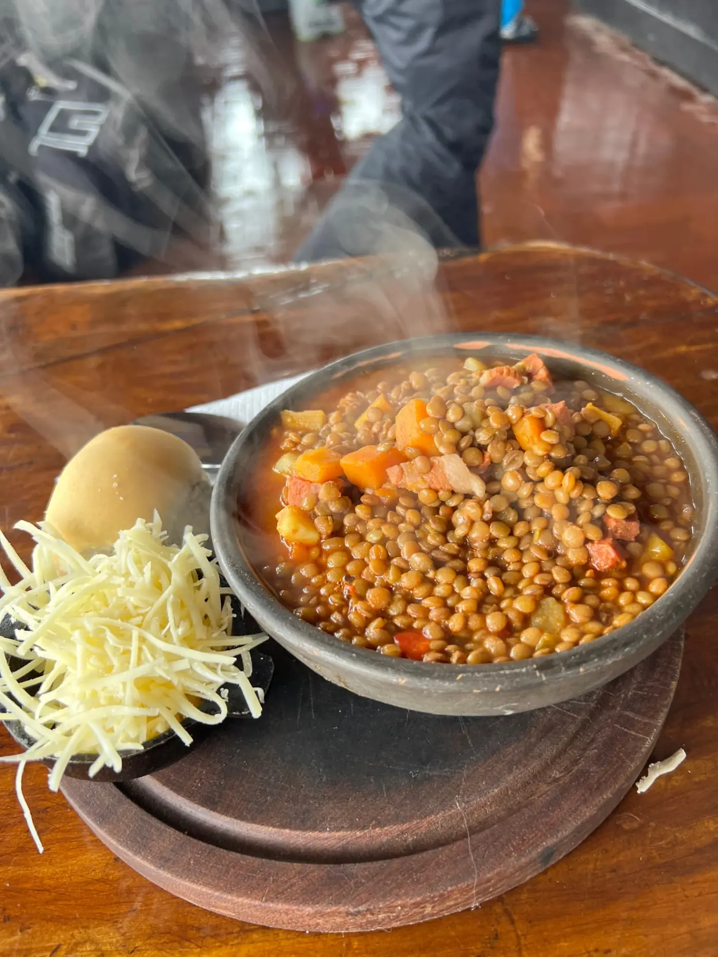 A steaming bowl of lentil stew with carrots and diced meat on a wooden board, served with a bread roll and a small dish of grated cheese