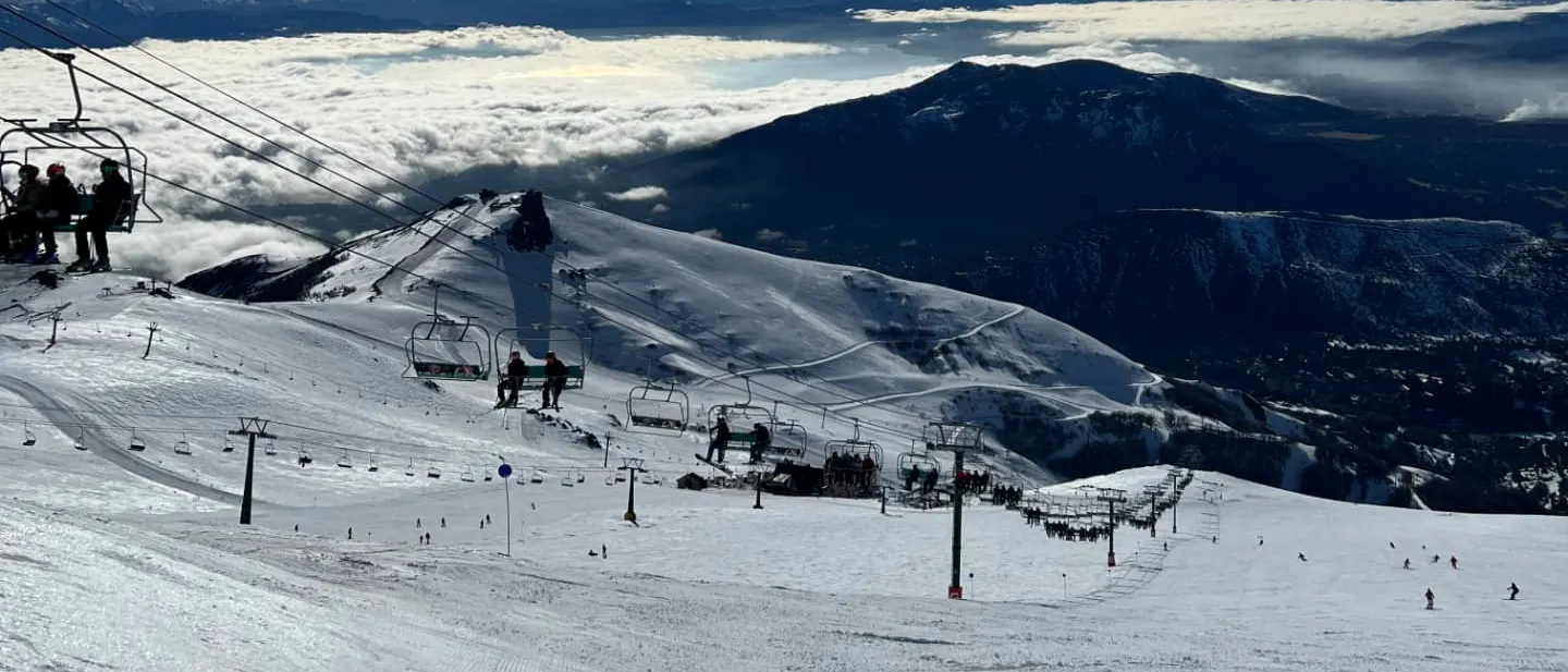 Skiers riding a chairlift at Cerro Catedral above a sea of clouds, with snow covered ridges and Patagonian mountains below