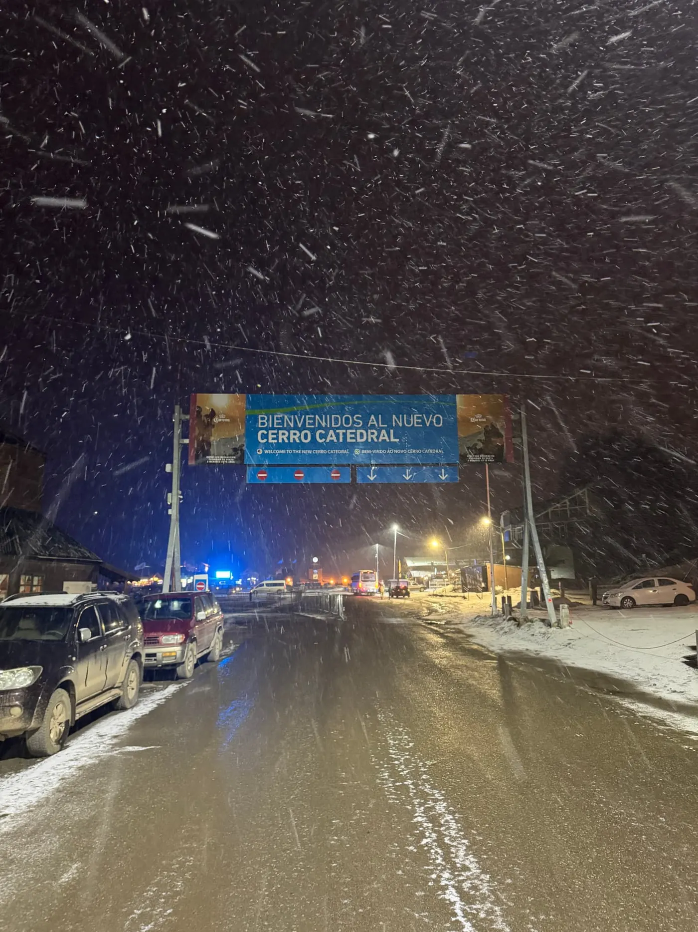 Cars and buses on the snow covered access road under the Cerro Catedral welcome sign during heavy night snowfall
